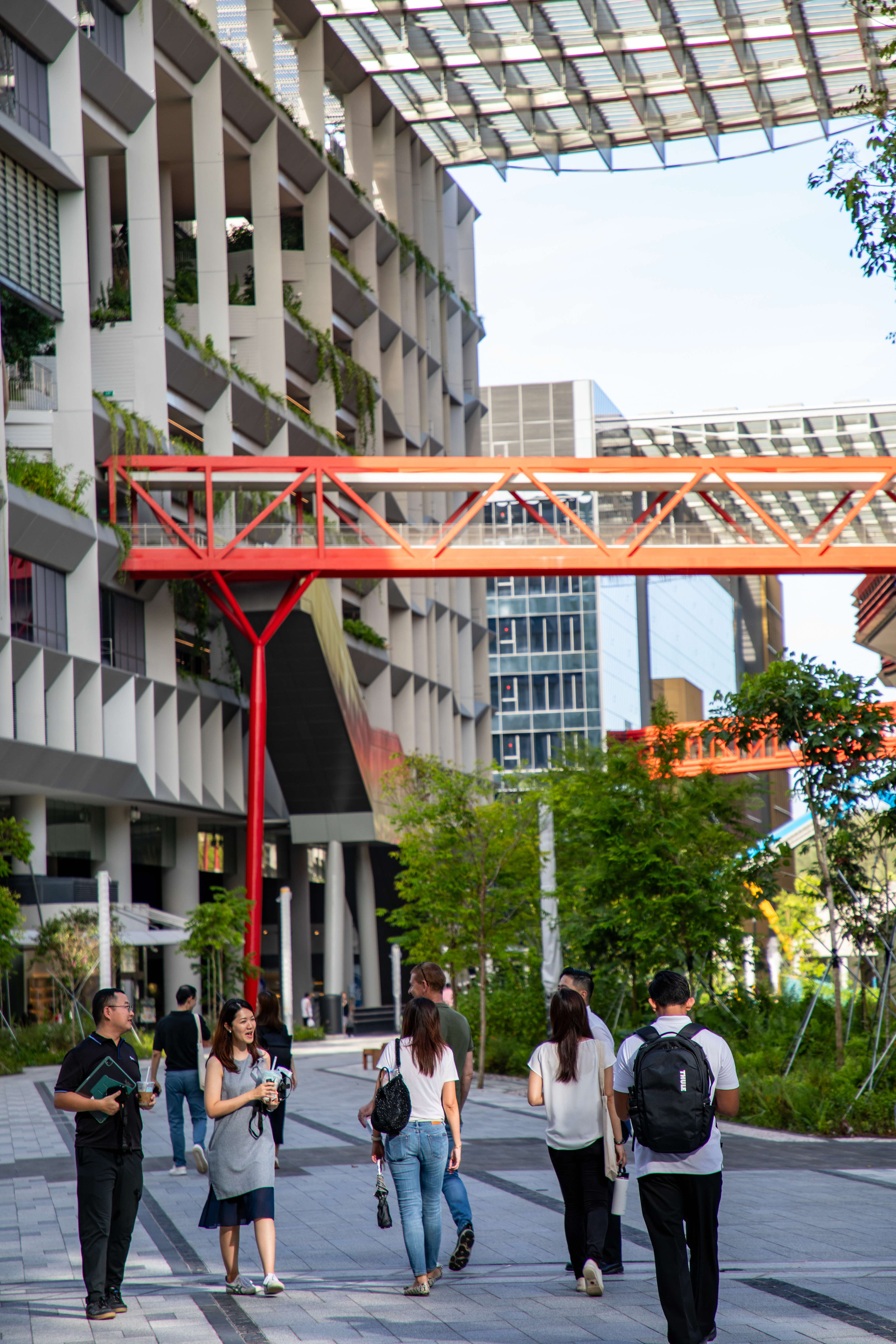 People walking on a plaza near modern buildings with a red skywalk.